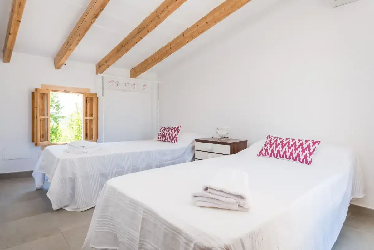 A bright bedroom with two beds, exposed wooden ceiling beams, and an open window.