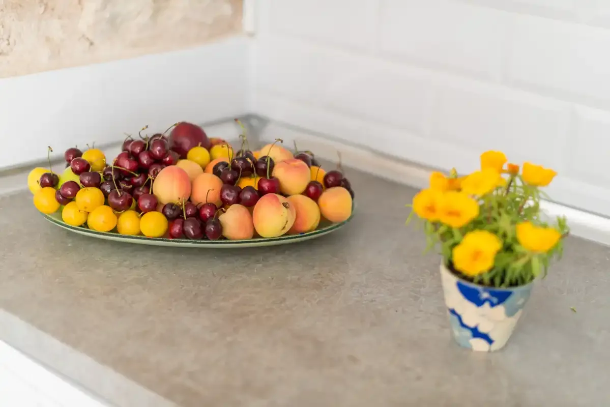 A kitchen counter holds a plate of assorted fruit beside a small vase of yellow flowers.