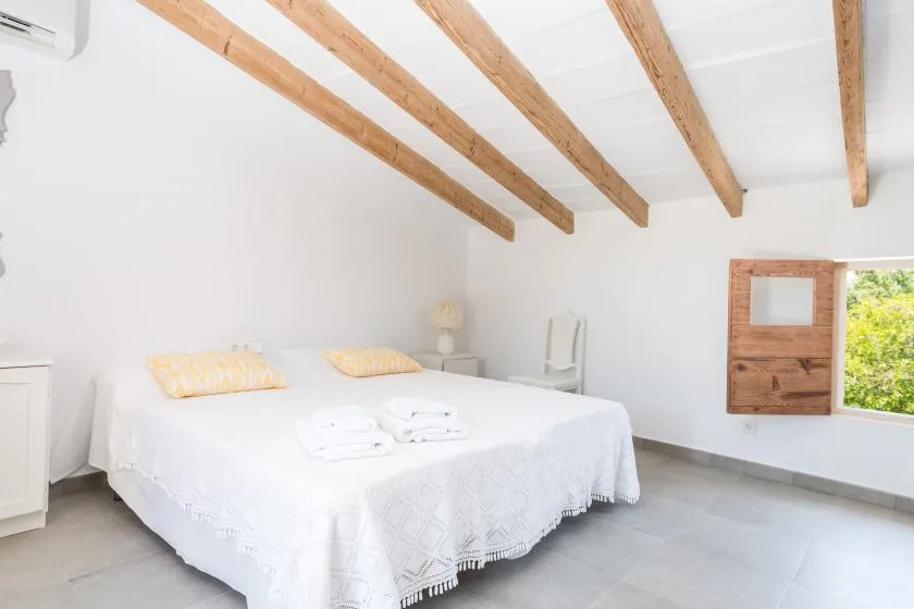 A bright bedroom with a white bed beneath exposed wooden ceiling beams.