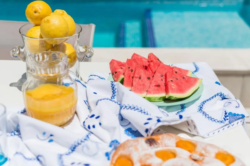 Fruit and pastries sit on a table beside a swimming pool.