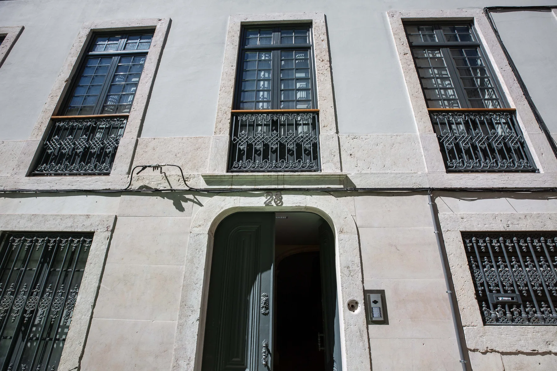 Una fachada de edificio clara tiene ventanas altas con rejas y una entrada abierta.