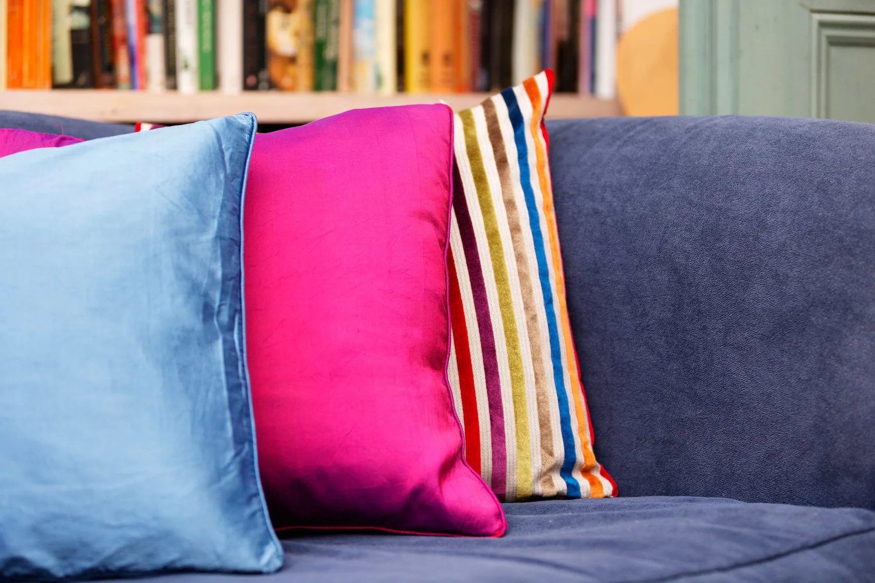 A blue couch with bright pink, blue, and striped cushions in front of a bookshelf.