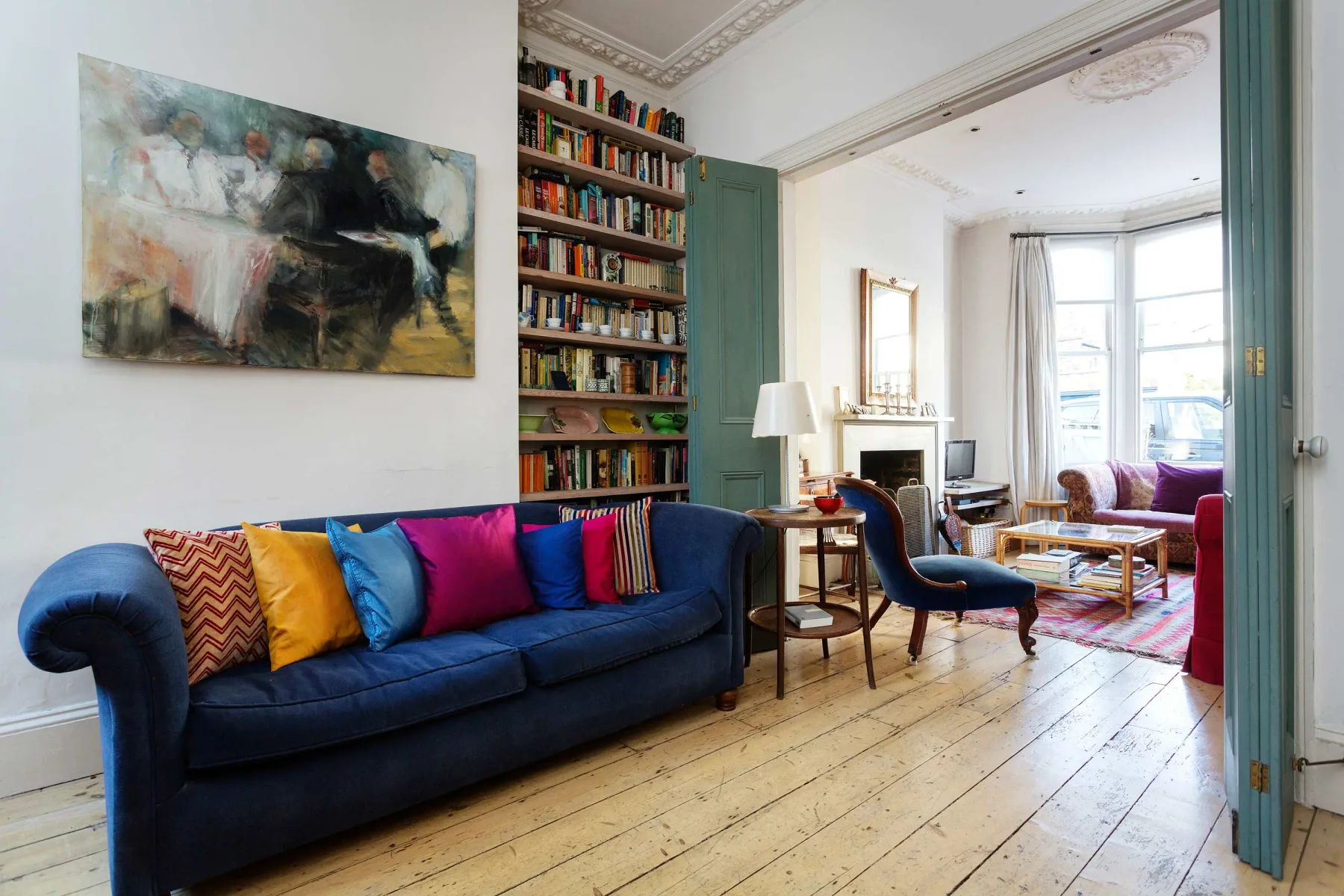 Una sala luminosa con un sofá azul, cojines de colores y una estantería alta.