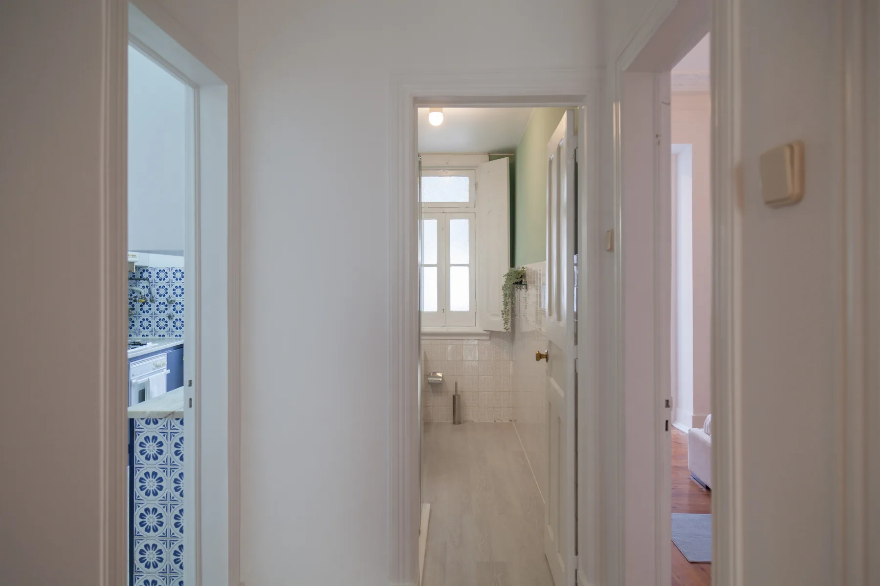 Un pasillo luminoso conecta una cocina con azulejos azules y blancos y un baño.