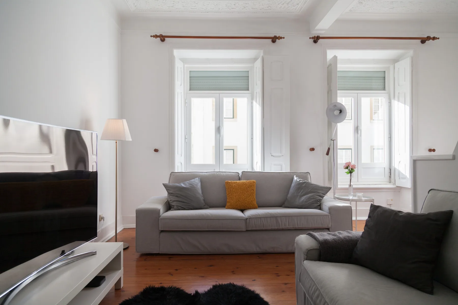 Sala de estar con asientos grises, dos ventanas altas y un televisor.