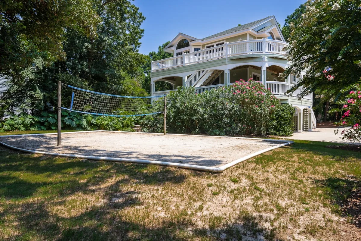 A sandy volleyball court sits in front of a large white house surrounded by trees.