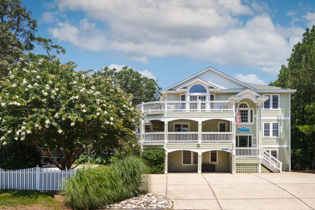 Una casa verde pálido con porches blancos está junto a árboles y una entrada pavimentada.