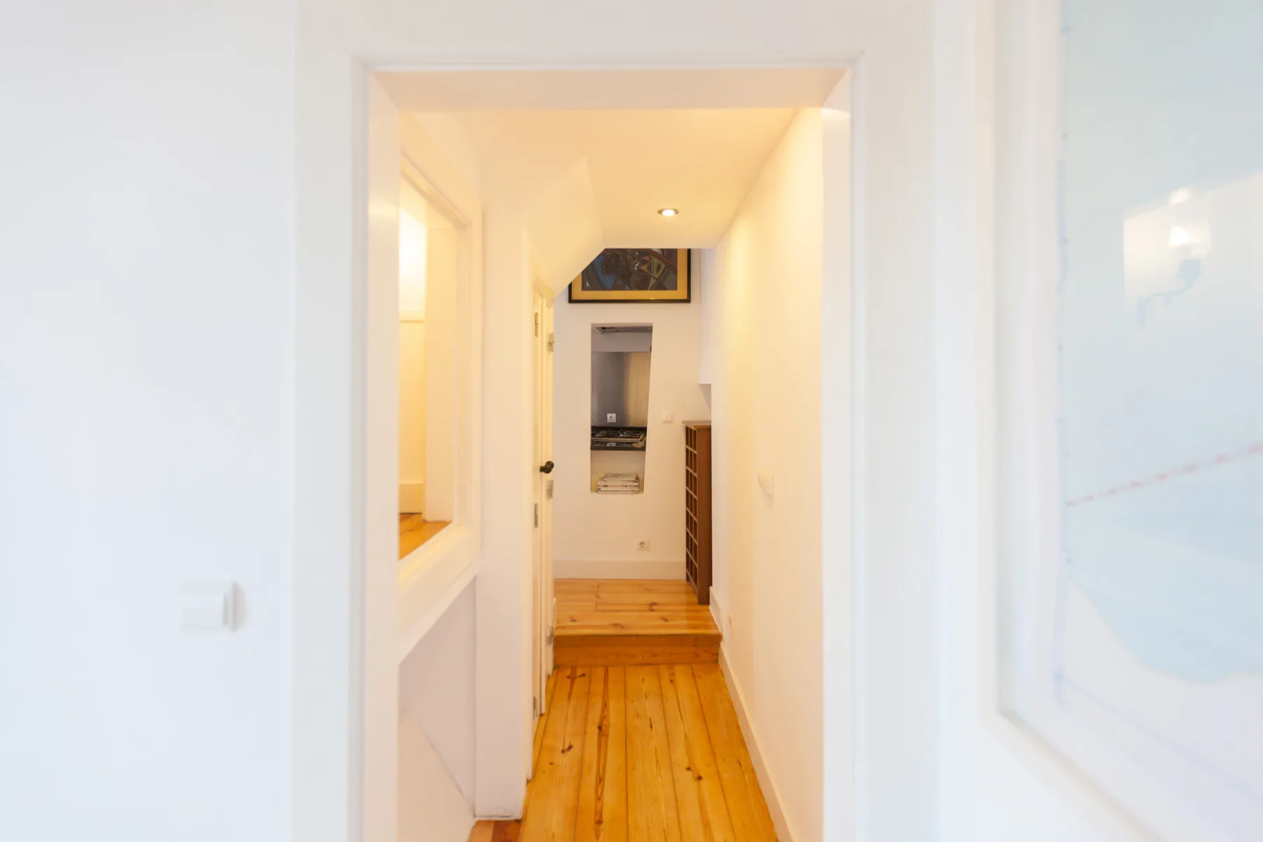 Un pasillo luminoso con suelo de madera conduce hacia una cocina.
