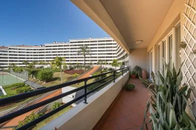 A balcony corridor with potted plants overlooks landscaped grounds and a large building.