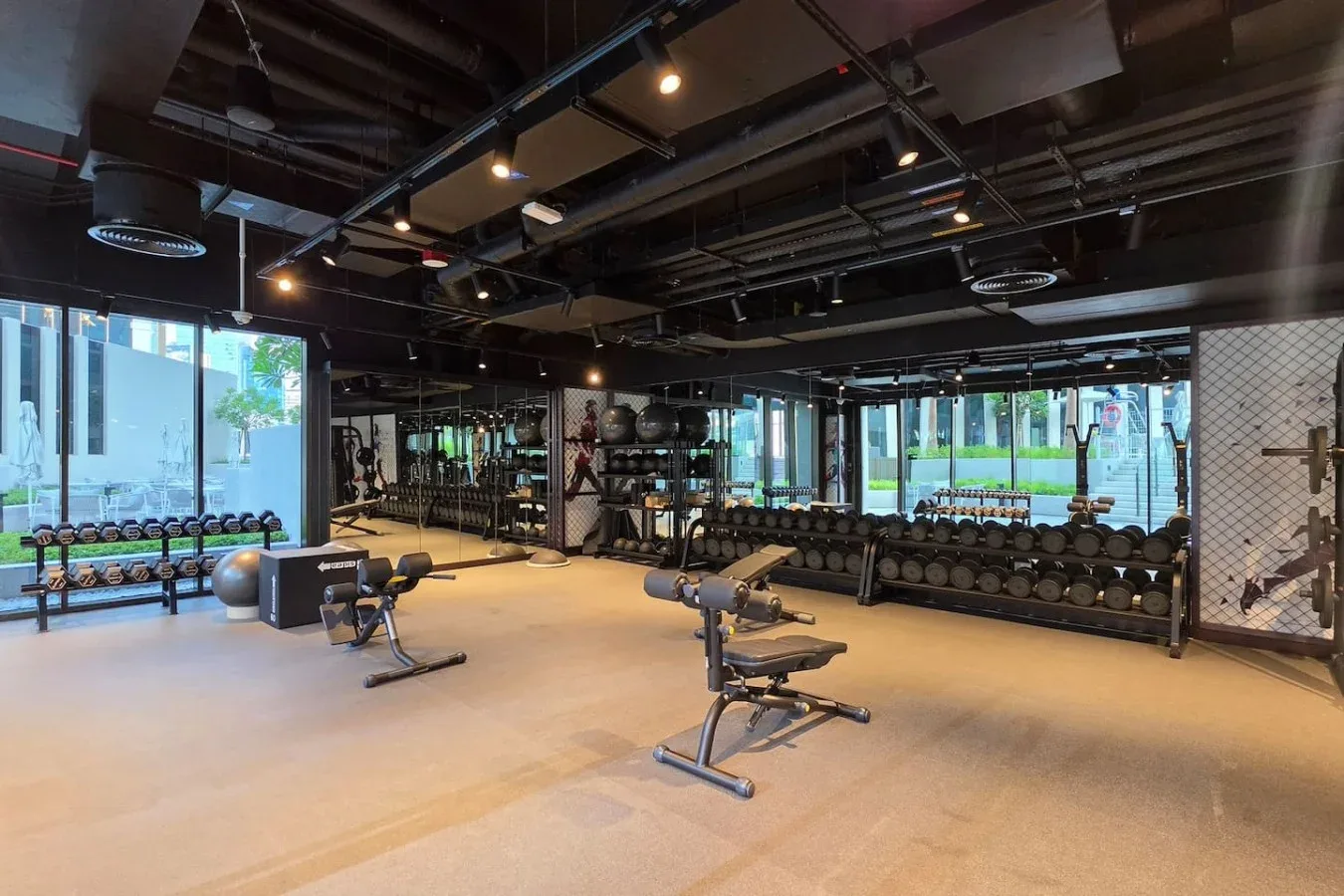 Un gimnasio amplio con estantes de pesas y bancos de ejercicio junto a grandes ventanas.