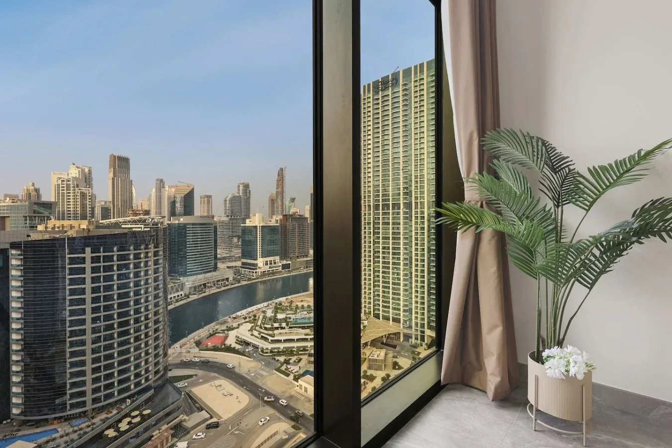 Se ven edificios altos y el perfil urbano por una ventana grande, junto a una planta en maceta.