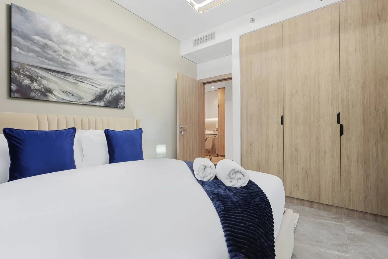 Un dormitorio con una cama tendida, cojines azules y armarios de madera clara.