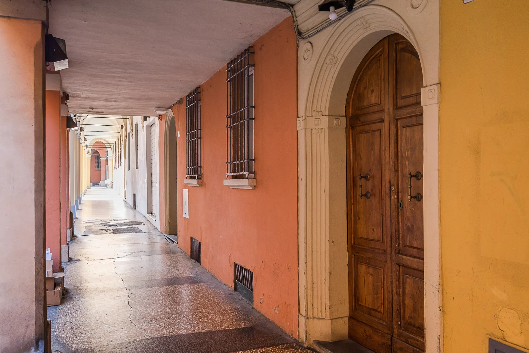 Un pasillo exterior cubierto pasa junto a paredes naranjas y una puerta arqueada de madera.