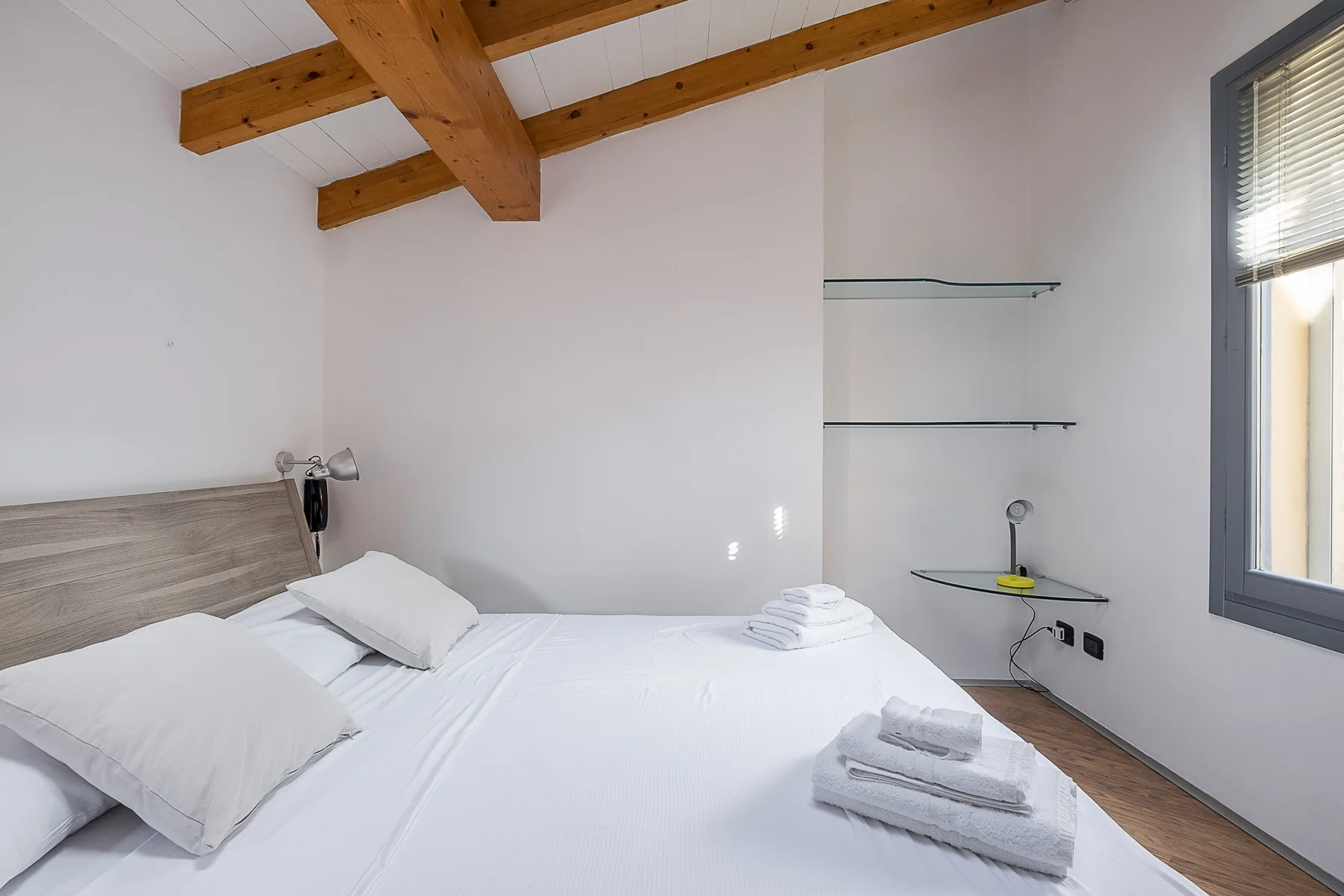 Un dormitorio con una cama blanca, vigas de madera a la vista y estantes de pared.
