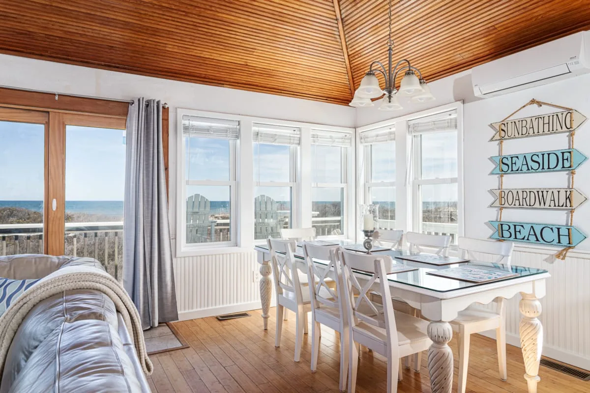 A bright dining area with a table and chairs beside windows overlooking the ocean.