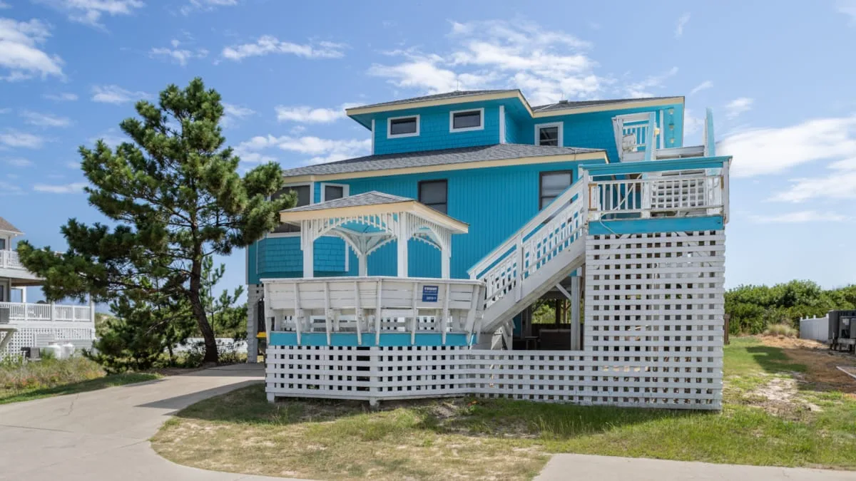 A bright turquoise raised house with white railings stands beside a paved drive.