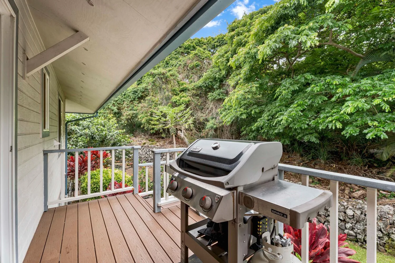 Una terraza cubierta tiene una parrilla junto a una barandilla blanca y vegetación frondosa.