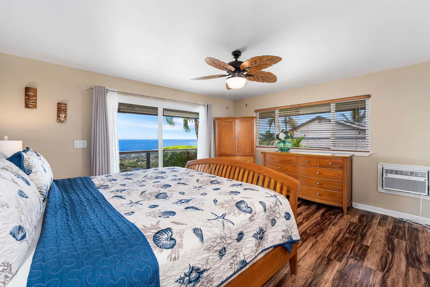 Un dormitorio con una cama estampada, una cómoda de madera y el océano visible por la ventana.