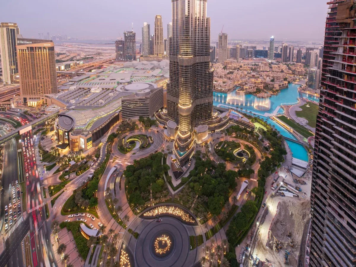 A city skyline overlooks a prominent tower, landscaped grounds, and a large turquoise pool.