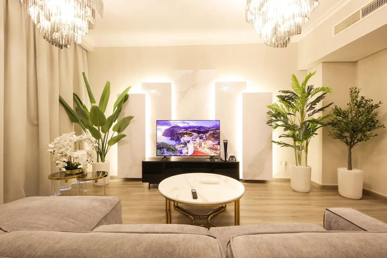 A bright living room features a television, a low table, and potted plants.