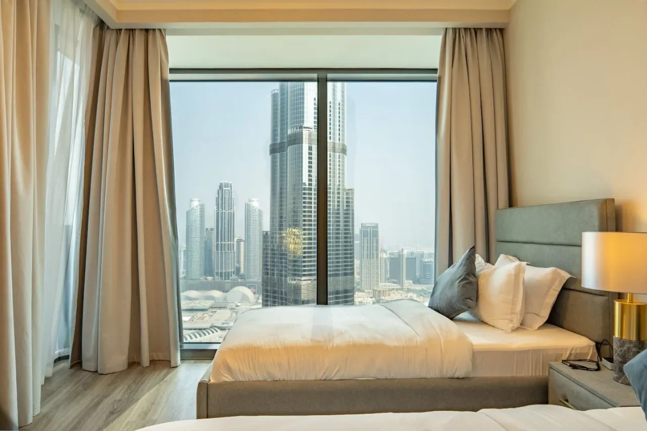 A bedroom with a bed beside a large window overlooking city skyscrapers.