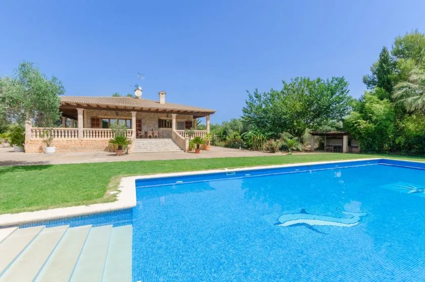 Una piscina ocupa el primer plano junto a un césped y una casa baja.