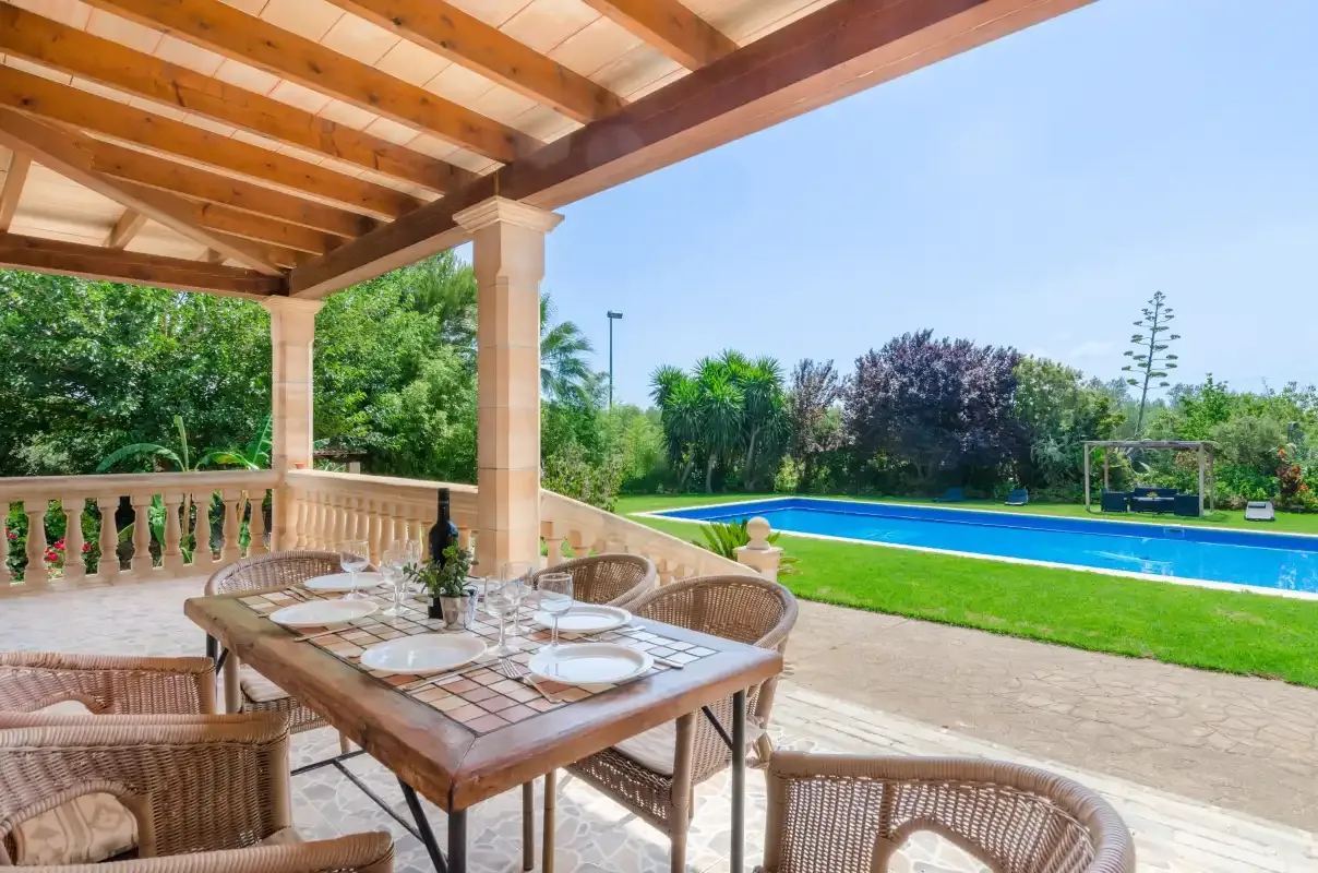 A covered patio with a dining table overlooks a swimming pool and garden.