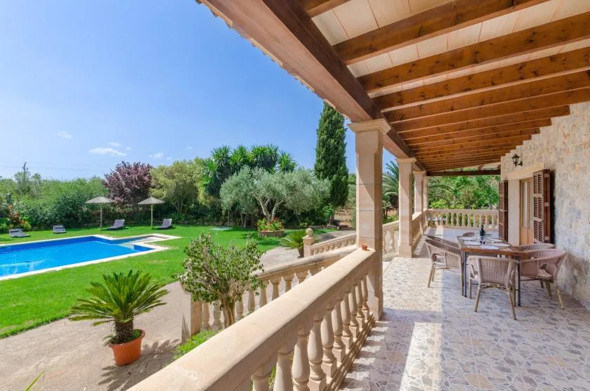 A covered stone terrace overlooks a lawn, garden, and swimming pool.