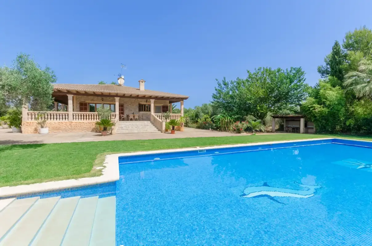 A swimming pool in front of a house, with green lawn and trees.