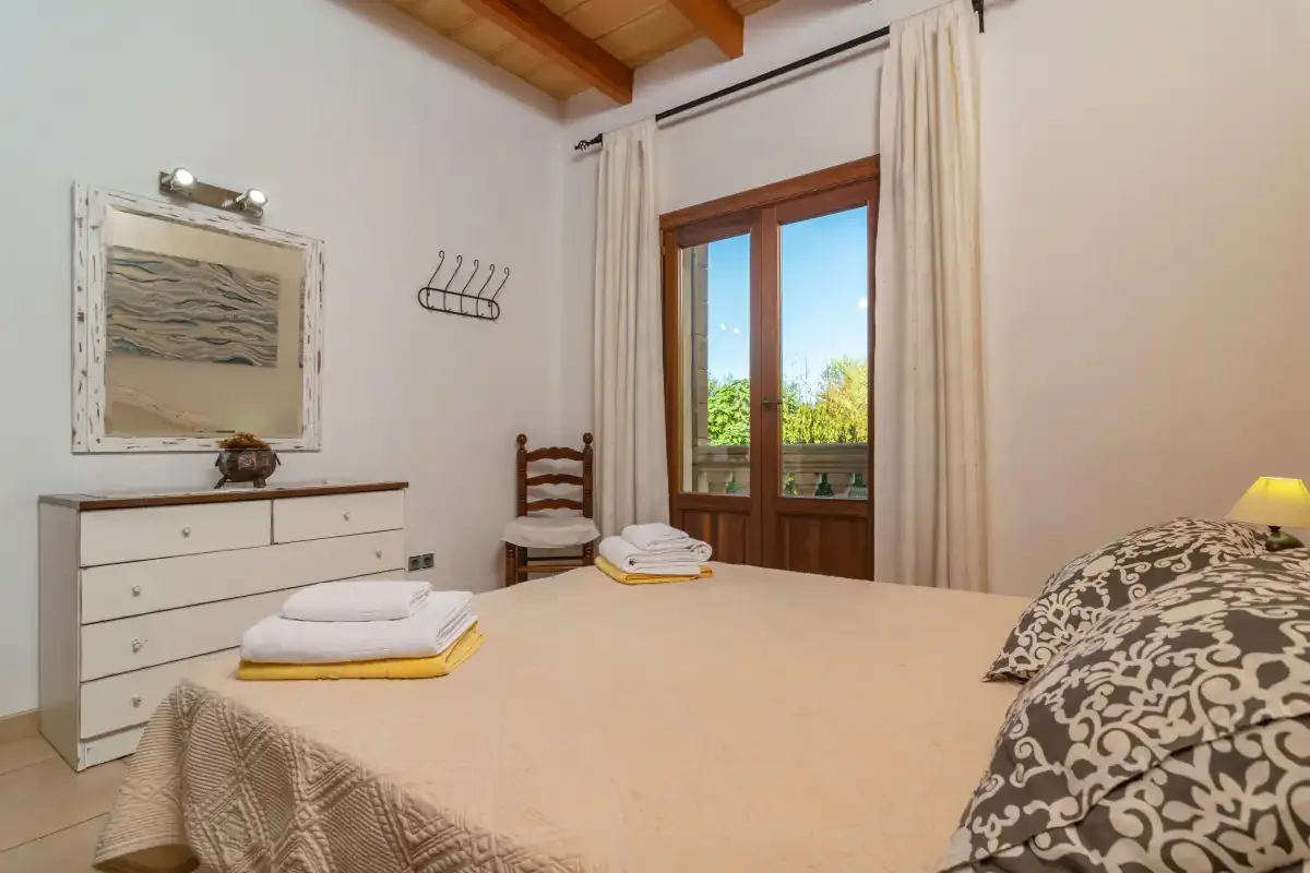 A bedroom with a neatly made bed, dresser, and wooden doors opening to a balcony.