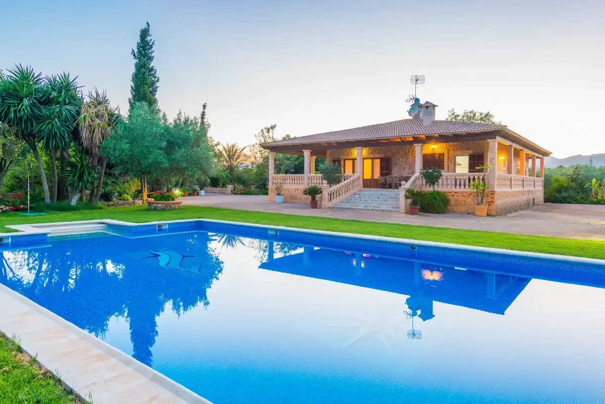 A house with a covered patio stands beside a swimming pool and landscaped garden.