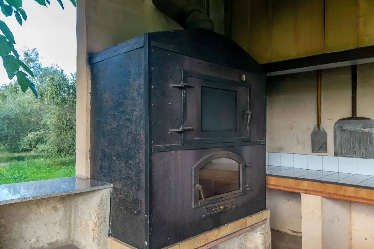 A large dark oven sits beside a tiled counter, with greenery visible through the open window.