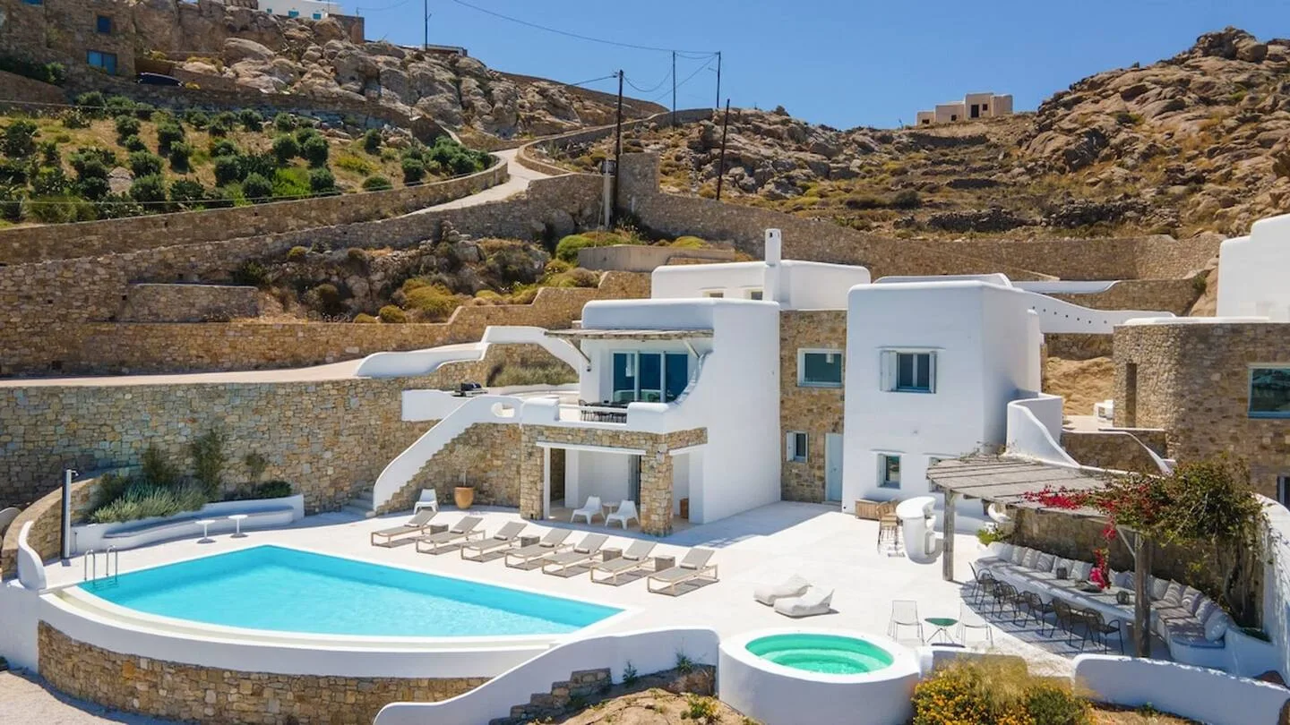 A modern white villa sits beside a swimming pool in a rocky hillside setting.