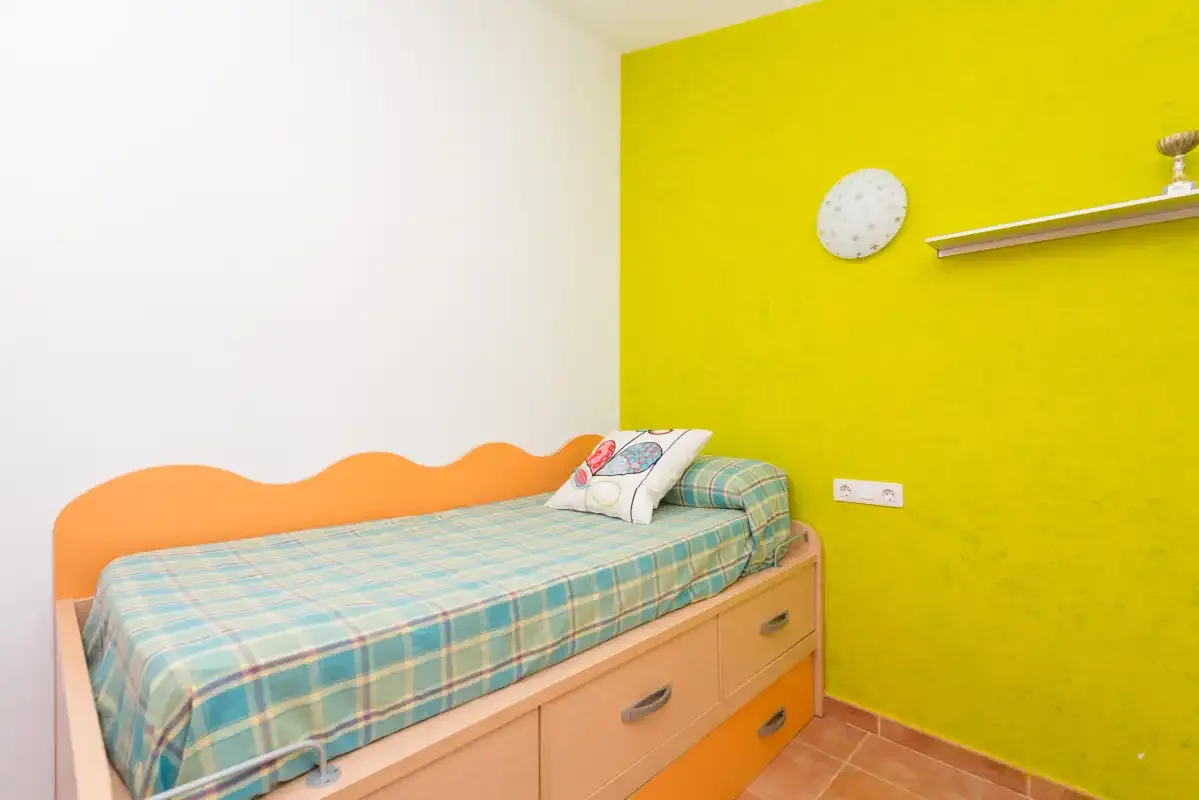 Una cama individual con ropa de cuadros está junto a una pared amarilla brillante.
