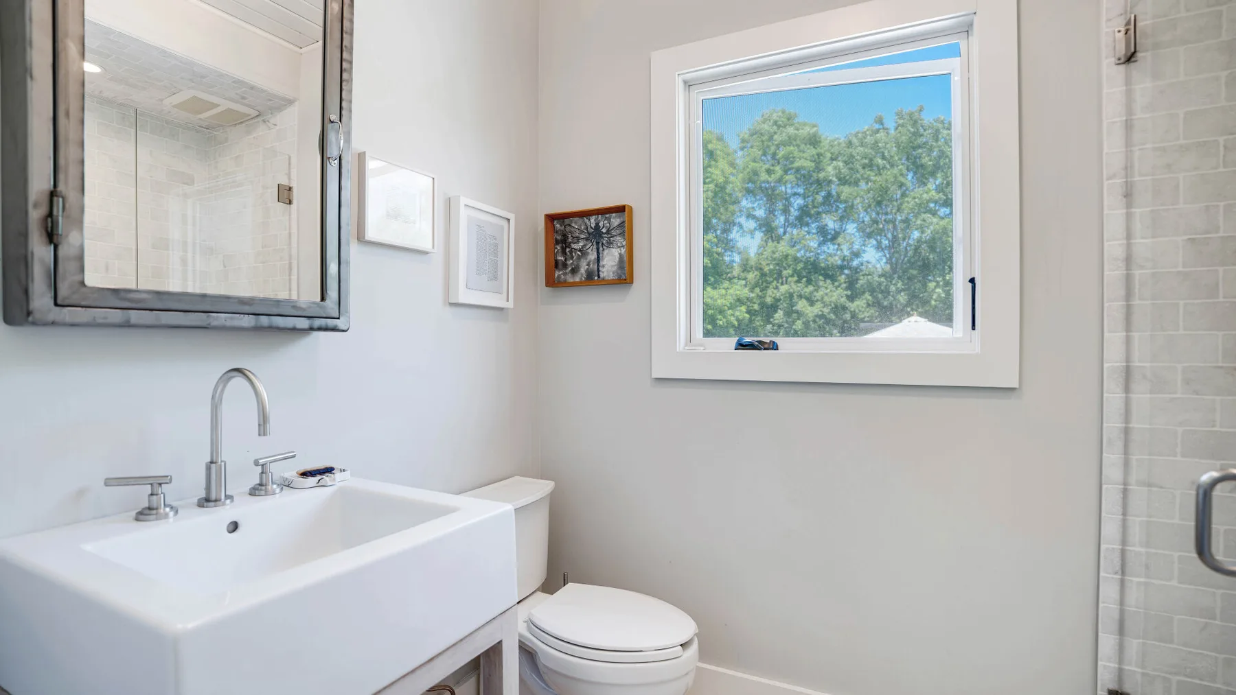 Un baño luminoso con lavabo, inodoro y una ventana con vista a árboles frondosos.