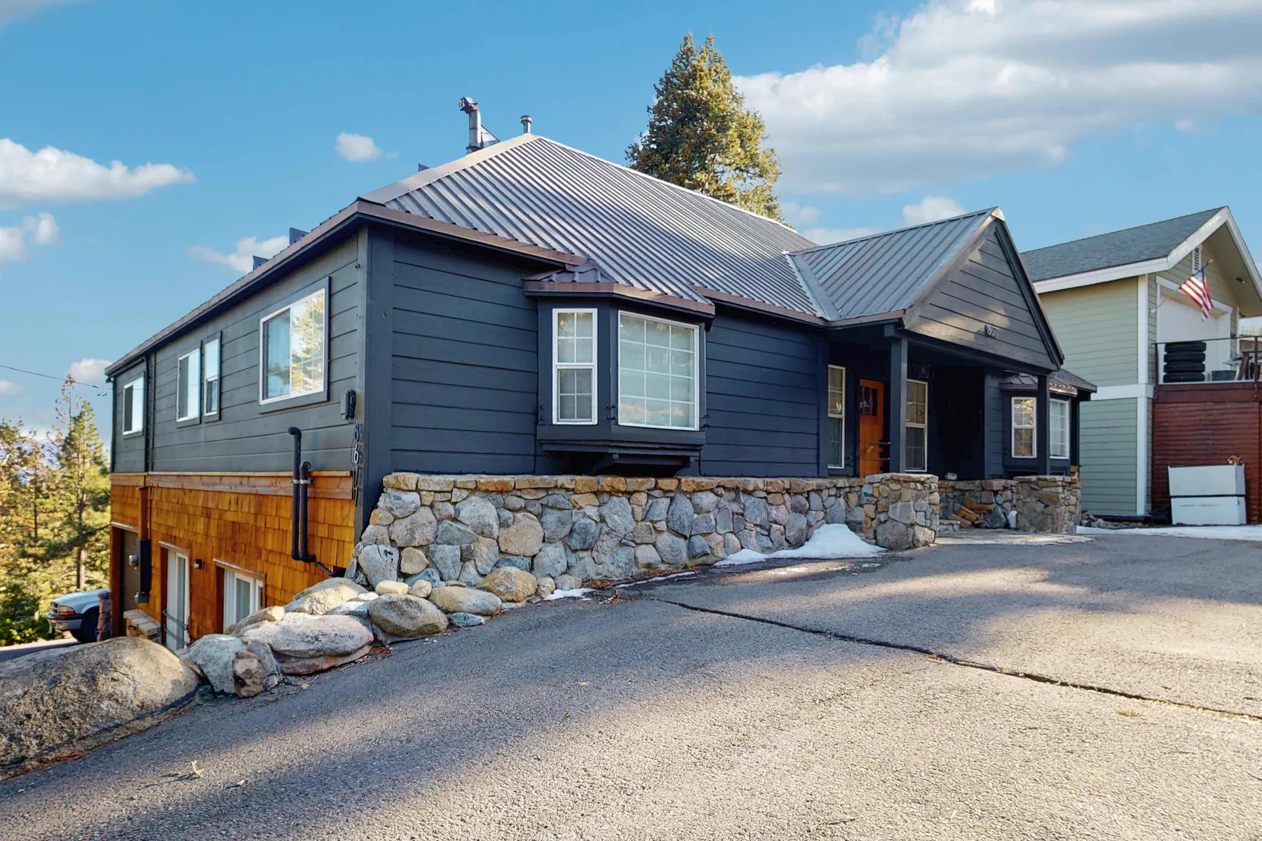 A dark-sided house with a stone foundation sits beside a paved driveway.