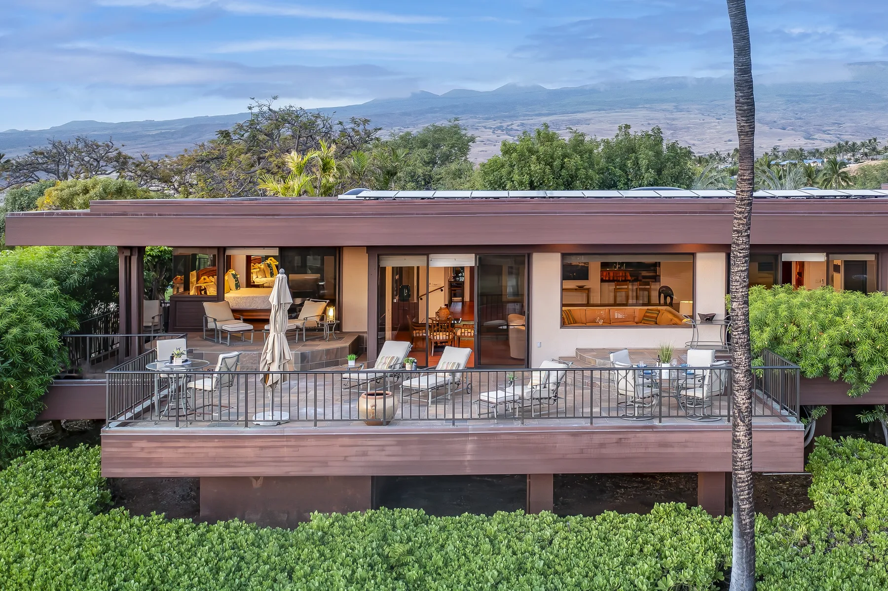 A modern house has a wide balcony overlooking green vegetation and distant hills.
