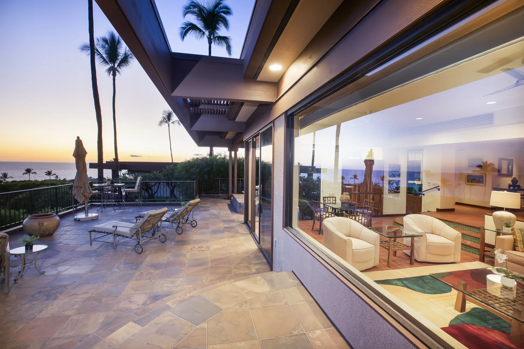 A furnished patio beside a glass-walled living area overlooks the coast at sunset.