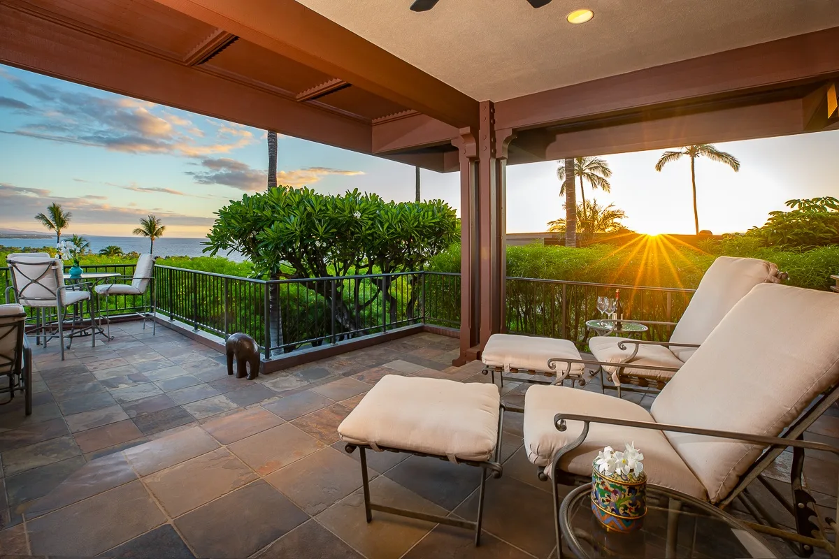 A covered outdoor patio with cushioned seating overlooks greenery and a sunset.