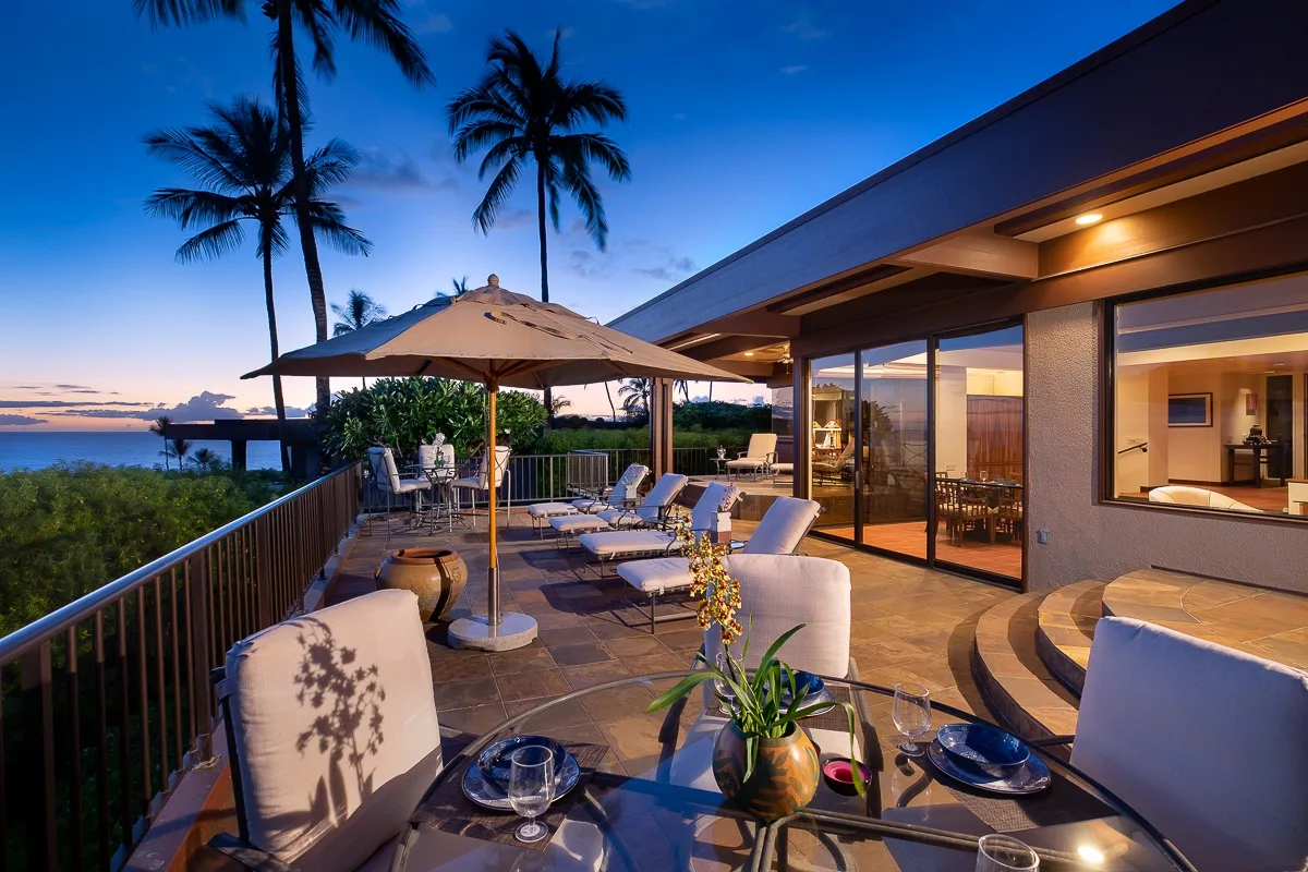A furnished outdoor terrace overlooks the water beside a modern house at dusk.