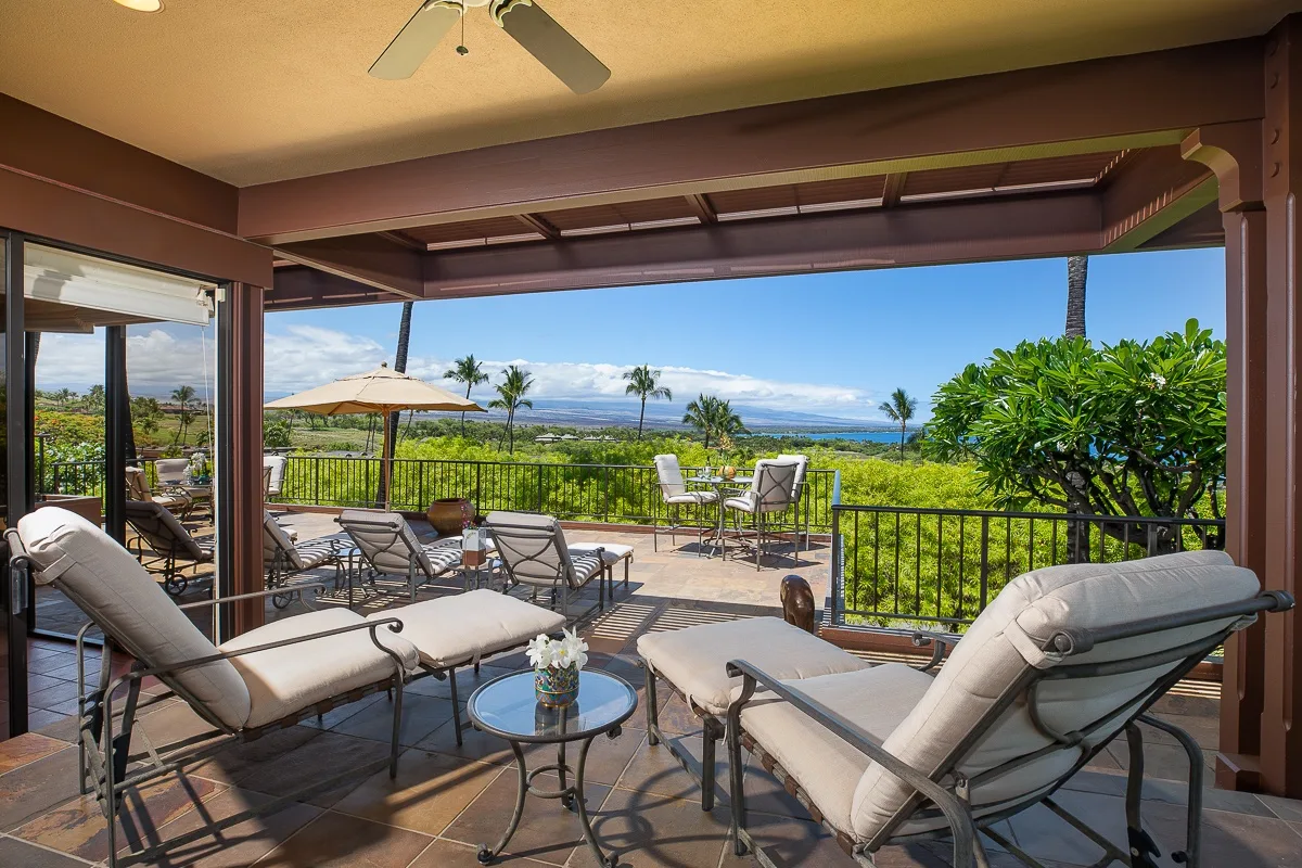 A covered outdoor patio with lounge seating overlooks a green landscape and distant ocean.