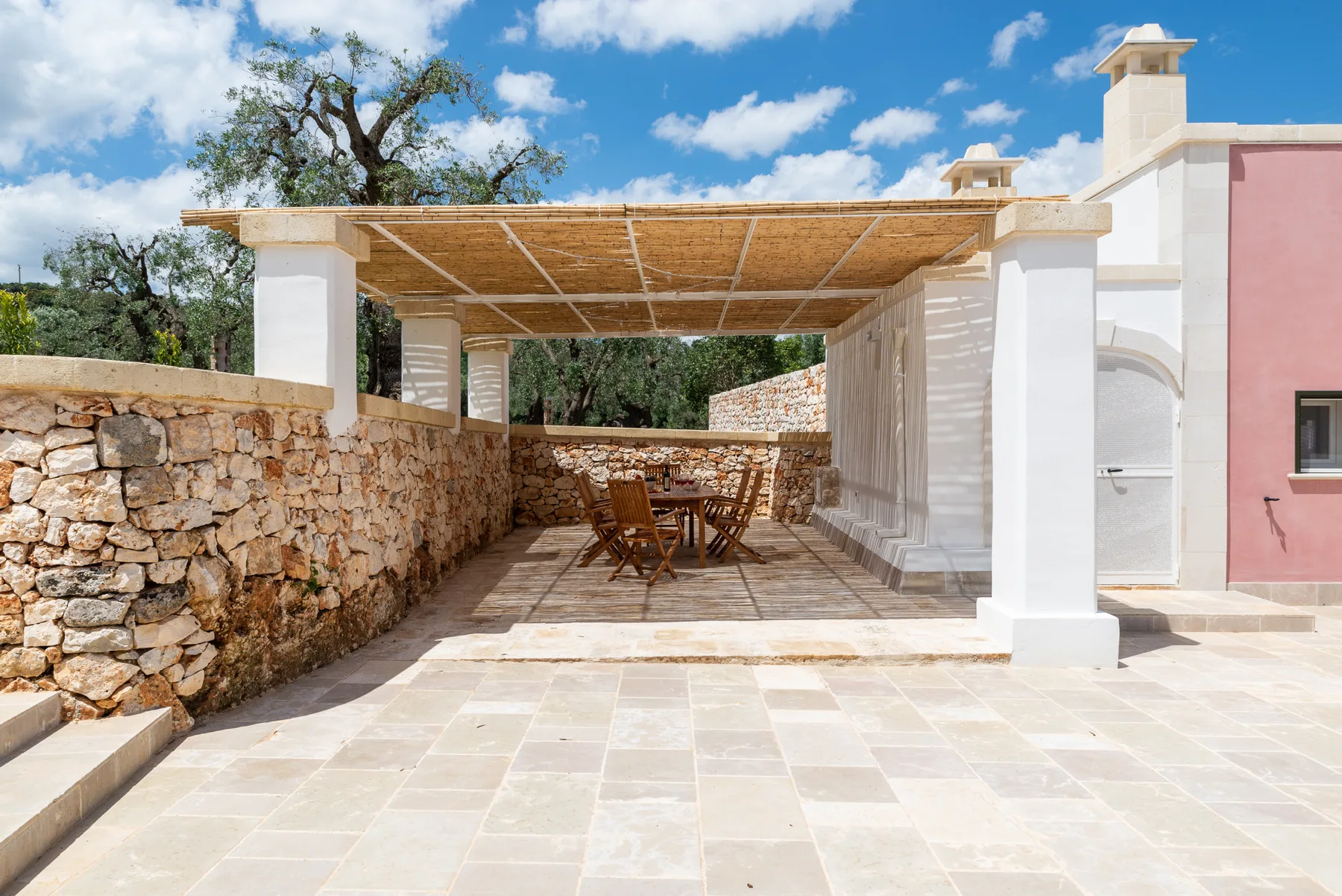 A paved outdoor terrace has a shaded dining area beside a stone wall.