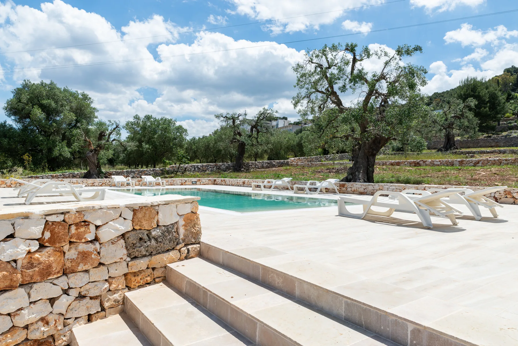 An outdoor swimming pool with pale loungers beside a stone wall.