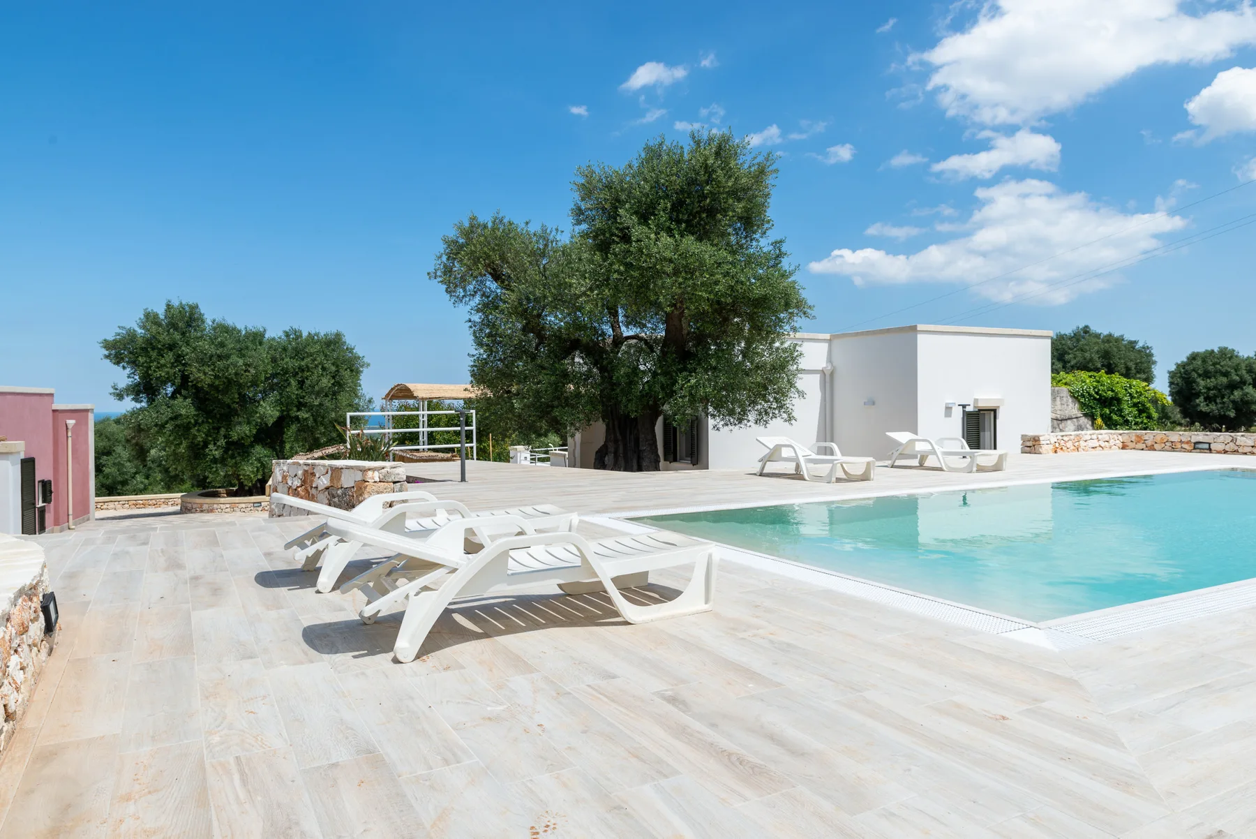 An outdoor swimming pool sits beside a paved deck with lounge seating and trees.