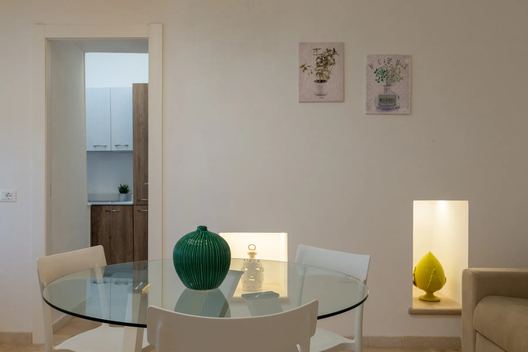A dining area with a glass table, green vase, and framed wall prints.