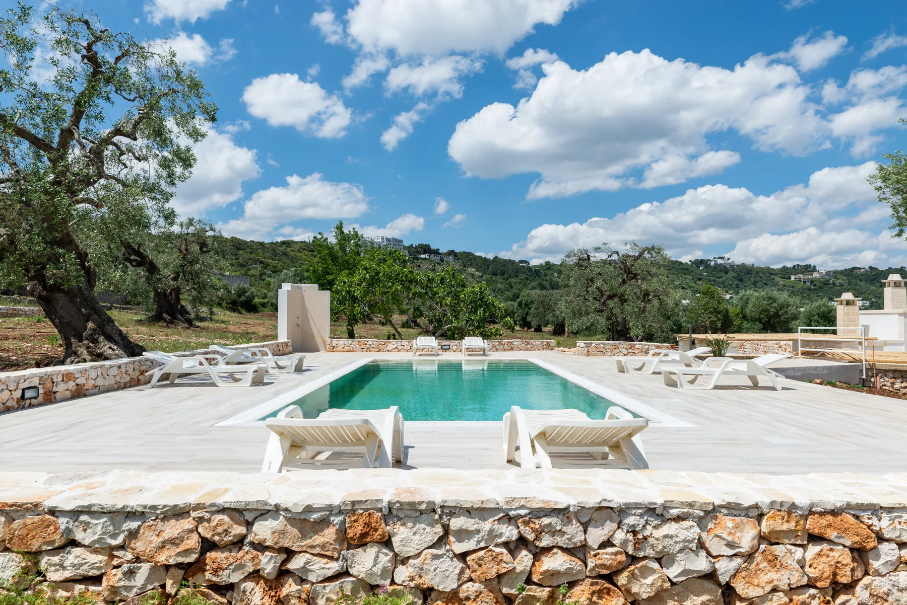 An outdoor swimming pool is surrounded by a pale stone deck and countryside.