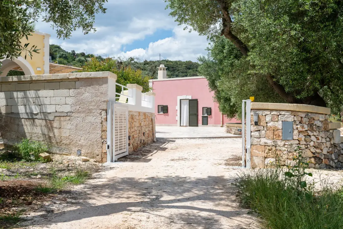 A sunlit stone entrance and path lead toward a pink building beneath leafy trees.