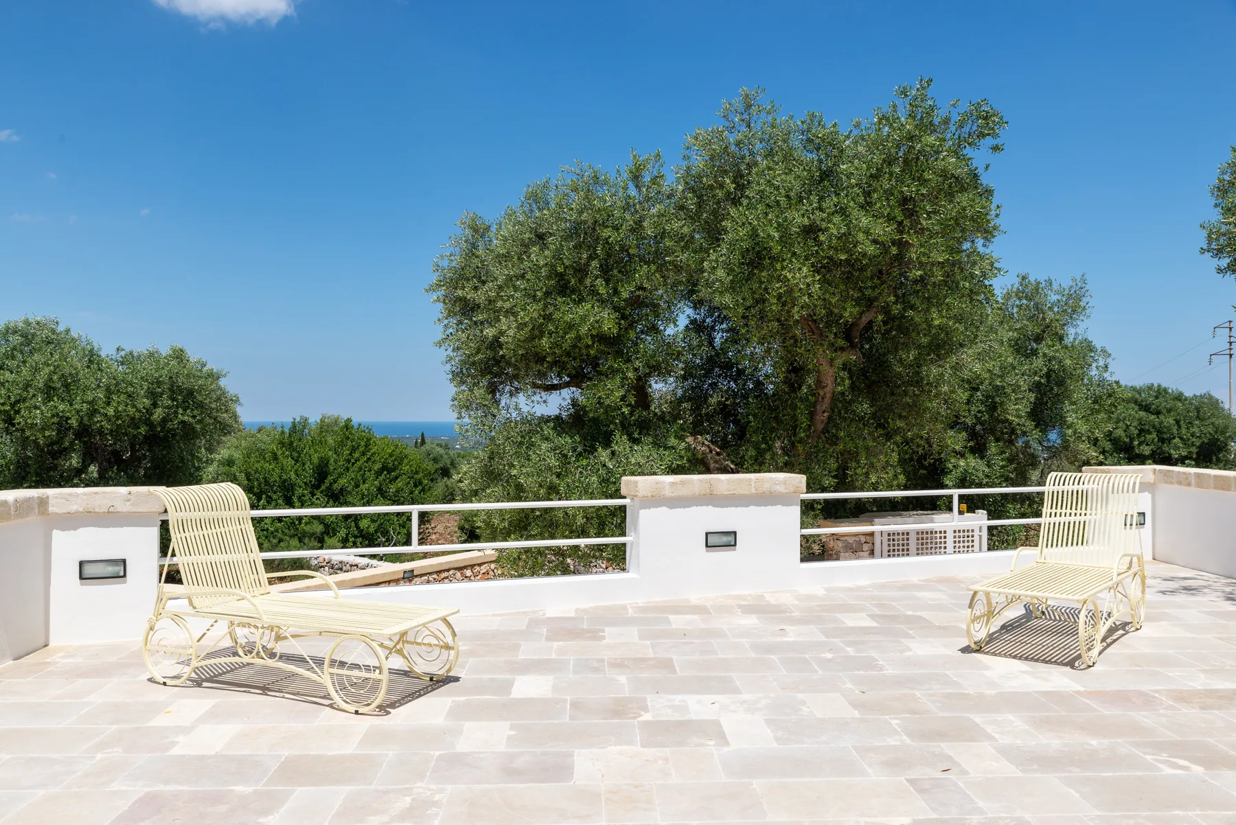 A sunlit outdoor terrace has pale paving, lounge seating, and leafy trees beyond the railing.