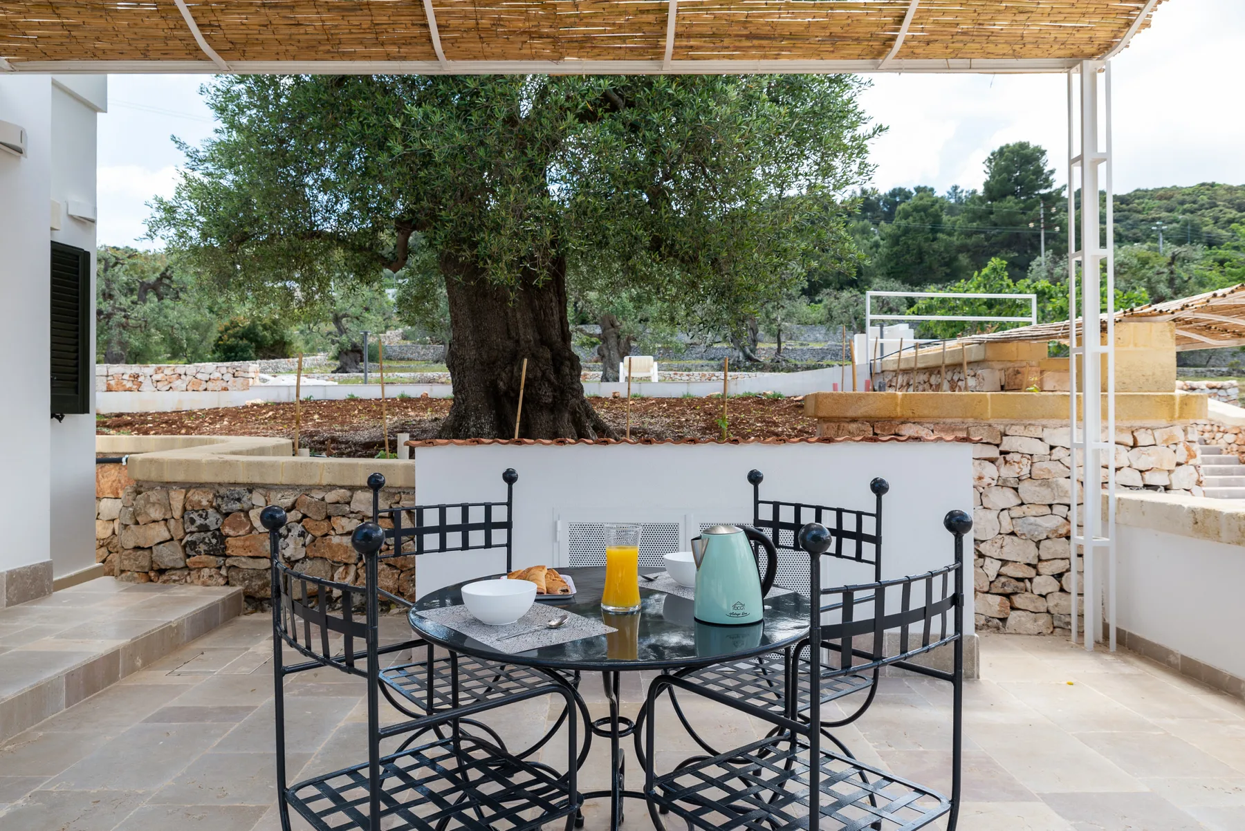 A covered outdoor dining area has a metal table and chairs beside a large tree.