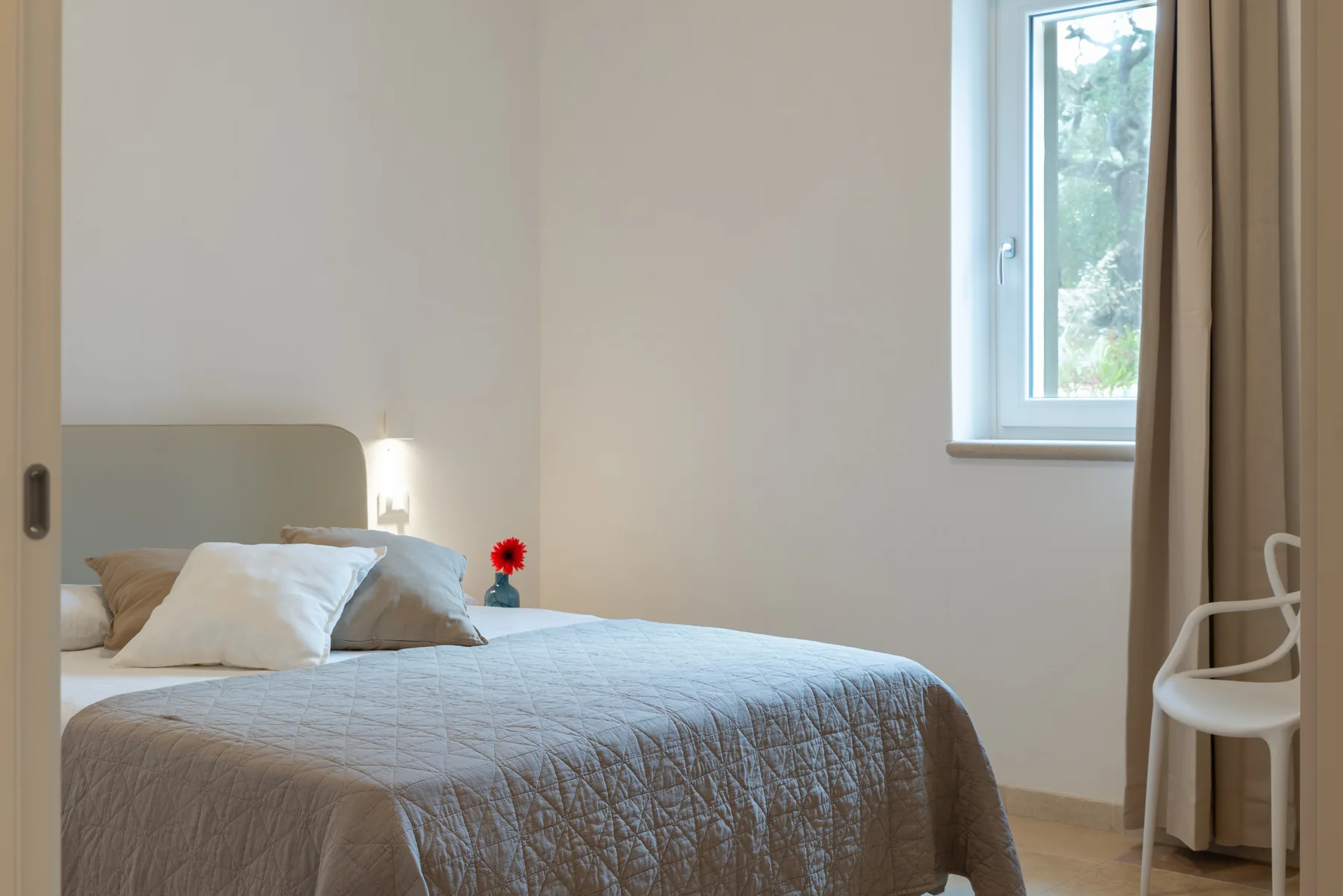 A bedroom with a bed, pale bedding, and a window beside the curtains.